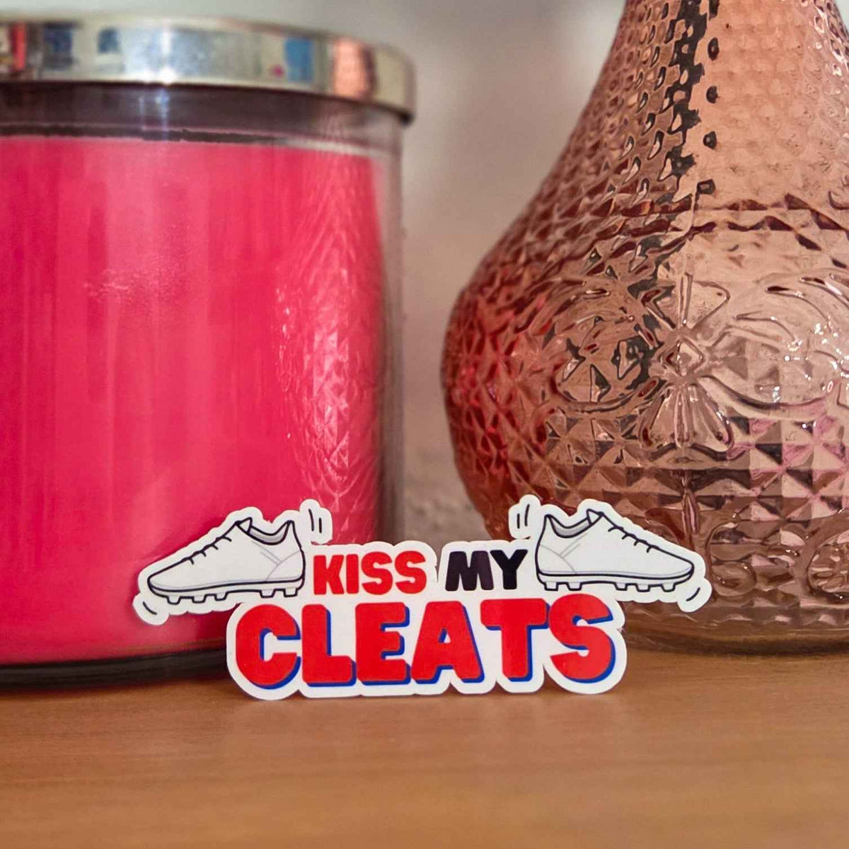 Pink candle and decorative vase with 'Kiss My Cleats' sticker on a wooden surface