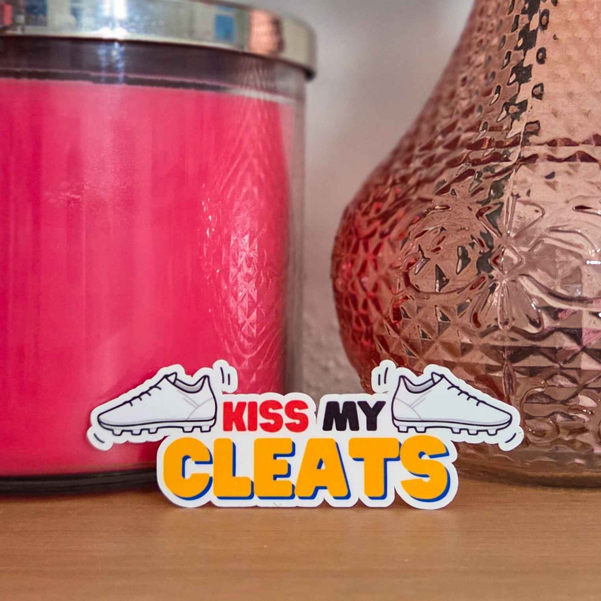Pink candle and decorative vase with 'Kiss My Cleats' sticker on a wooden surface