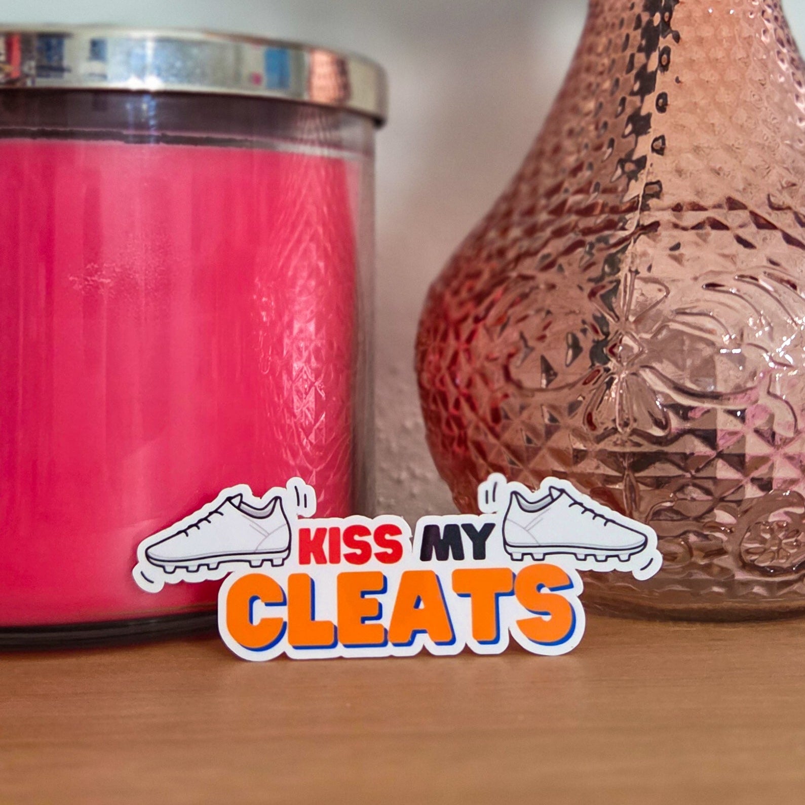 Pink candle and textured vase with a pink rose on a wooden surface, with 'Kiss My Cleats' logo.