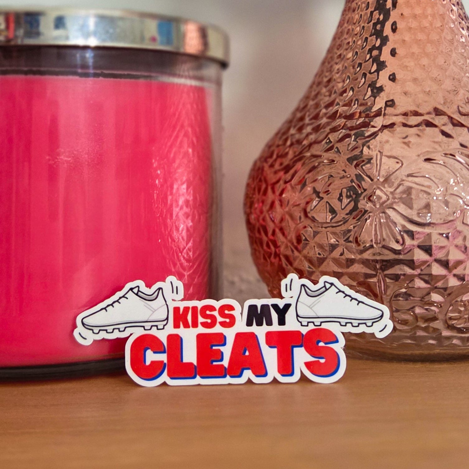 Pink candle and textured vase with a rose on a wooden surface, featuring 'Kiss My Cleats' logo.