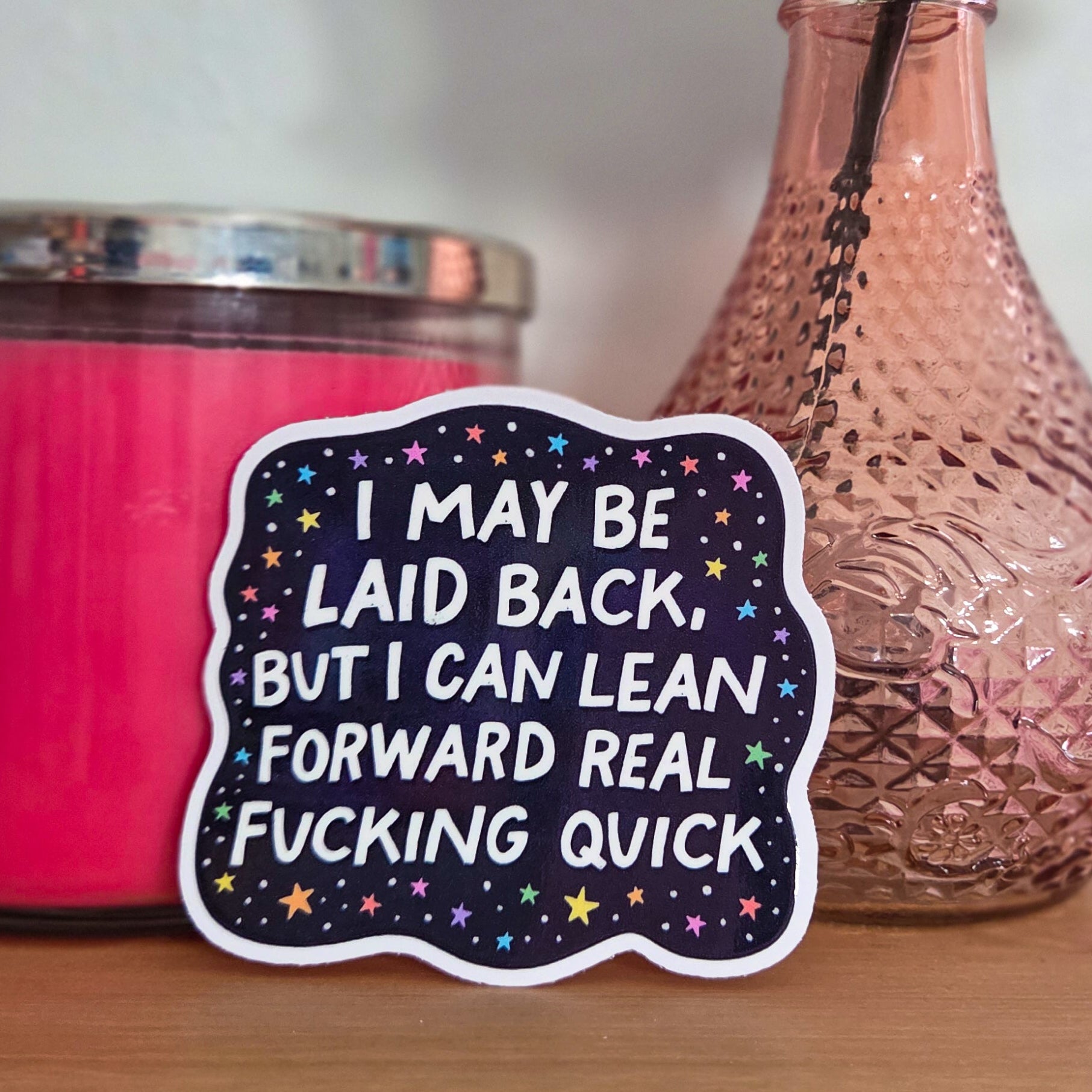 Sticker with humorous text on a wooden surface with a pink candle and vase with a rose.