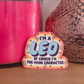 A die-cut sticker with a colorful astrological design that reads 'I'M A LEO OF COURSE I’M THE MAIN CHARACTER’  resting on a wooden surface.