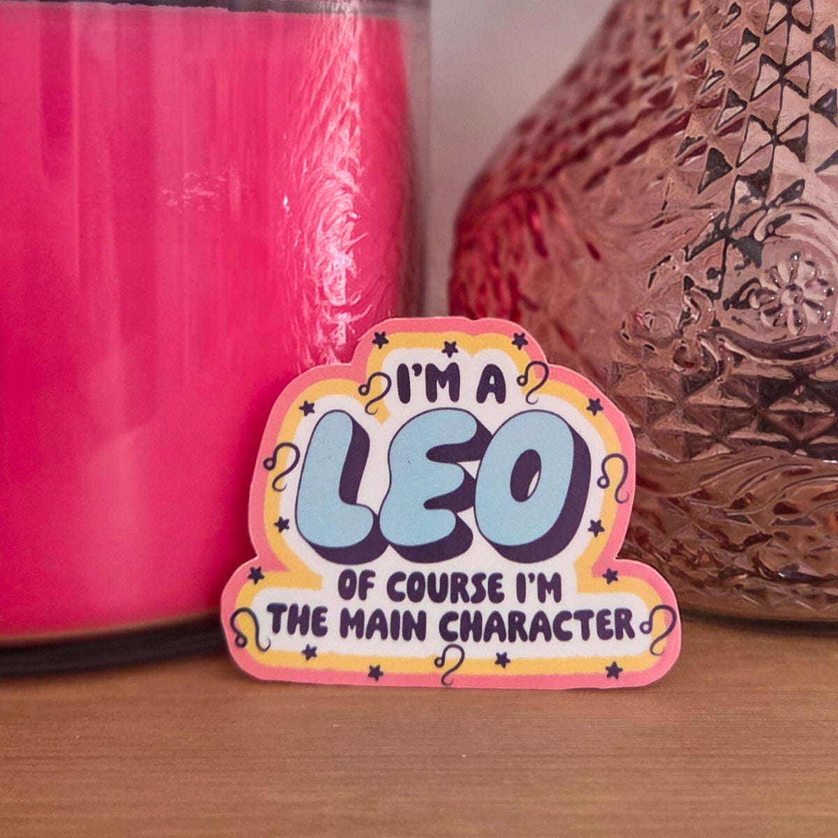 A die-cut sticker with a colorful astrological design that reads 'I'M A LEO OF COURSE I’M THE MAIN CHARACTER’  resting on a wooden surface.