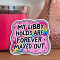 Sticker with text 'My Libby Holds Are Forever Maxed Out' on a pink background, next to a pink candle and vase with flowers.