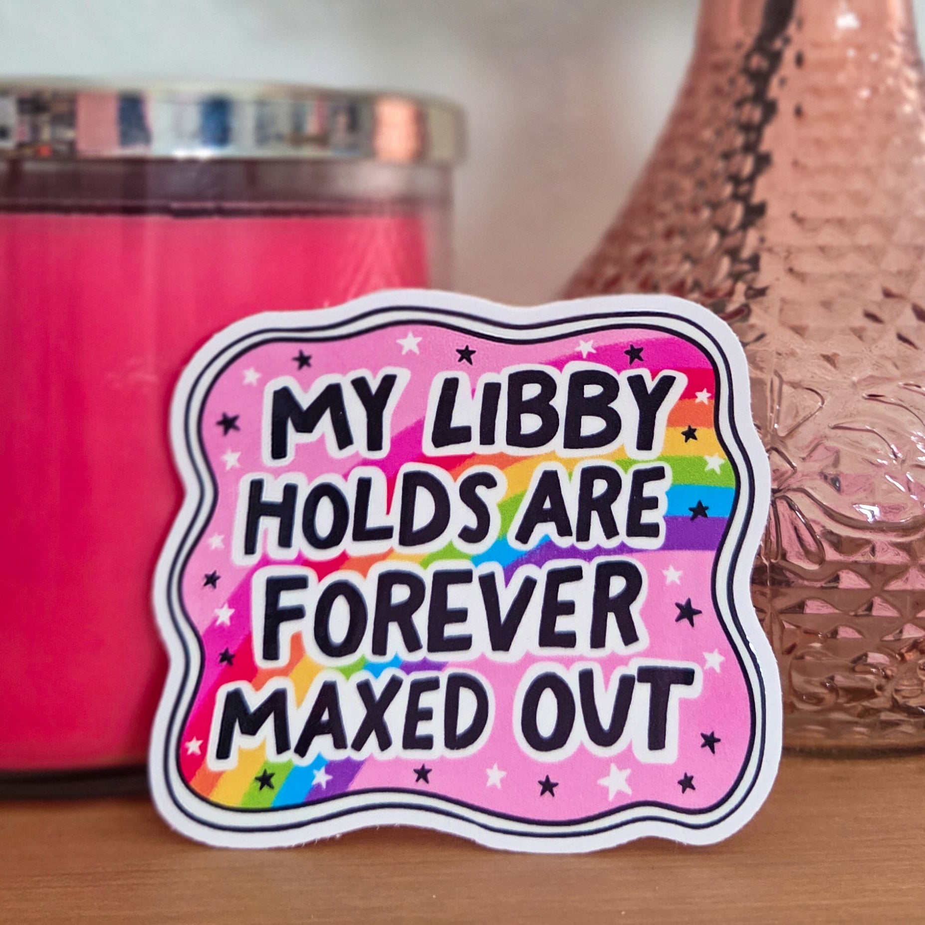 Sticker with text 'My Libby Holds Are Forever Maxed Out' on a pink background, next to a pink candle and vase with flowers.