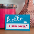 Sticker with 'hello I'm a Libby Lover' text next to a pink candle and vase with flowers on a wooden surface.