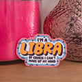 A die-cut sticker with the text 'I'M A LIBRA OF COURSE I CAN'T MAKE UP MY MIND' in yellow and black lettering, displayed on a wooden surface next to a pink candle and a textured brown glass vase.
