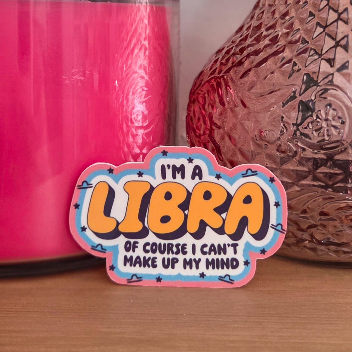 A die-cut sticker with the text 'I'M A LIBRA OF COURSE I CAN'T MAKE UP MY MIND' in yellow and black lettering, displayed on a wooden surface next to a pink candle and a textured brown glass vase.