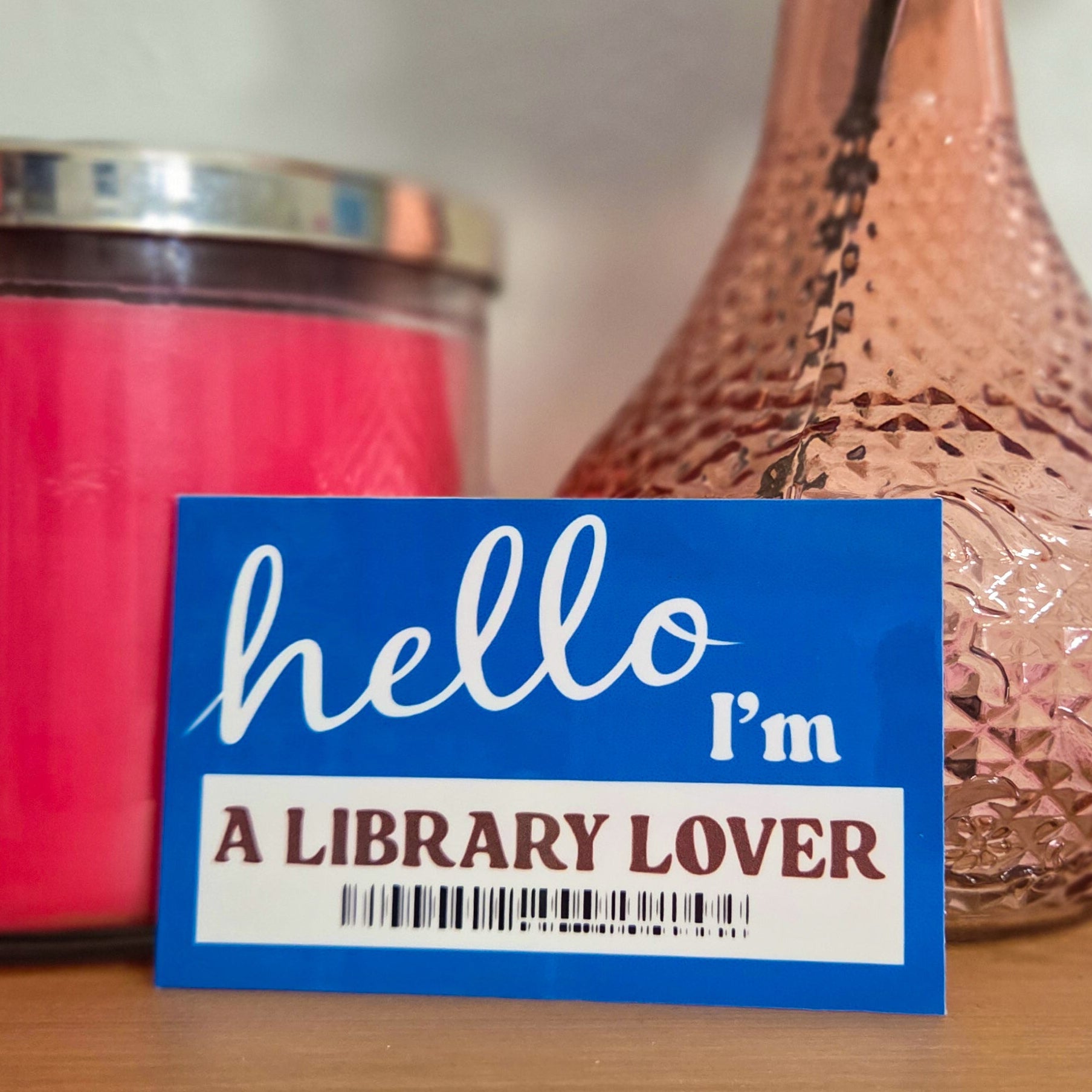 Blue card with 'hello I'm a library lover' text in front of a pink vase with a rose on a wooden surface.