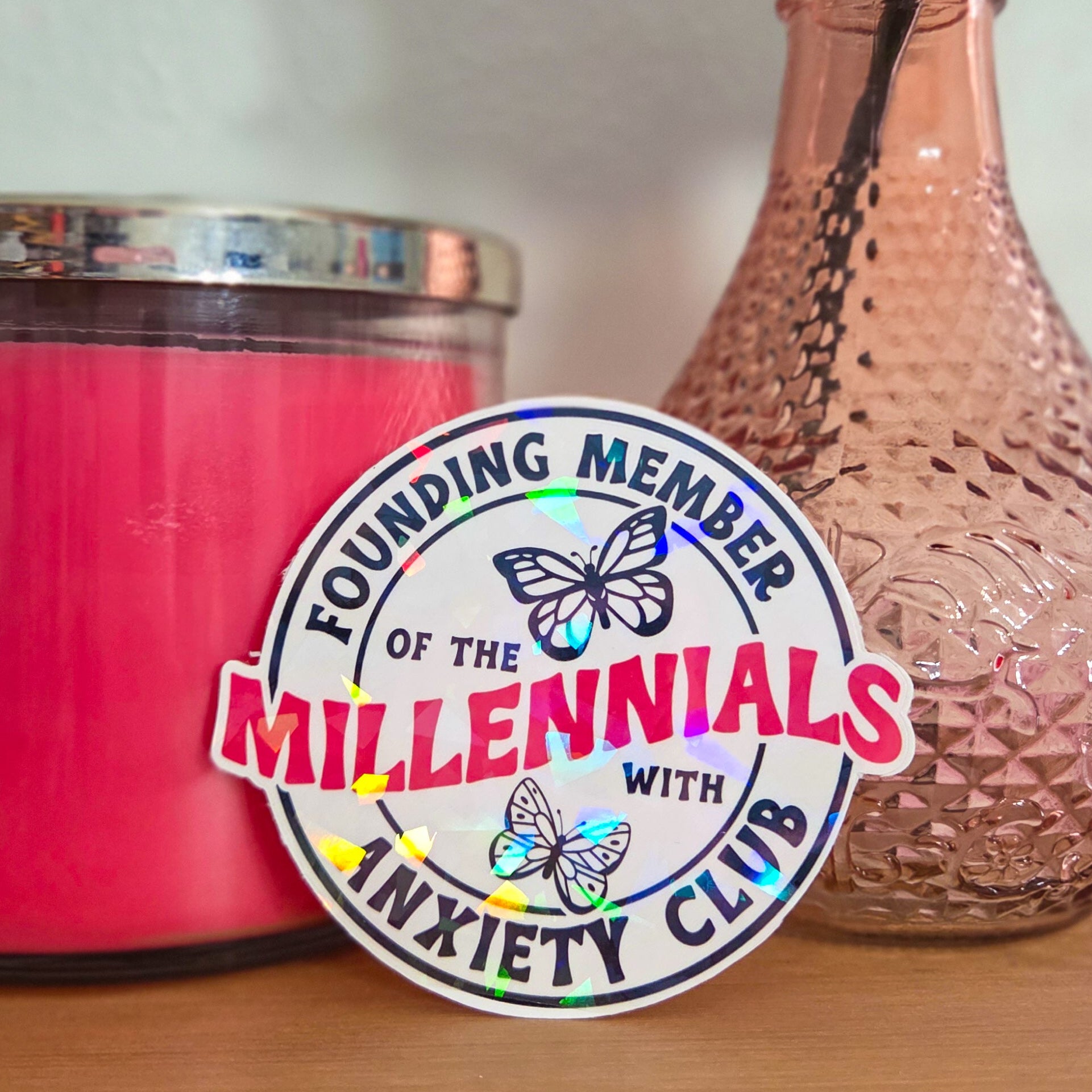 Pink candle and vase with a 'Founding Member of the Millennials with Anxiety Club' sticker on a wooden surface.
