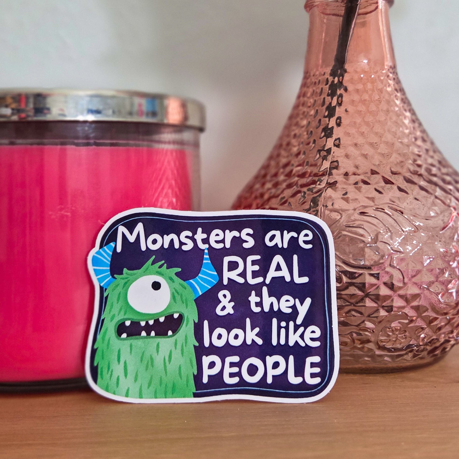 Decorative sticker with monster design next to a pink candle and vase with a flower on a wooden surface.