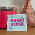 Small sticker with 'Moody Bitch' text next to a pink candle and vase with a rose on a wooden surface.
