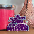 Pink candle, textured vase with a rose, and a humorous horoscope sticker on a wooden surface.