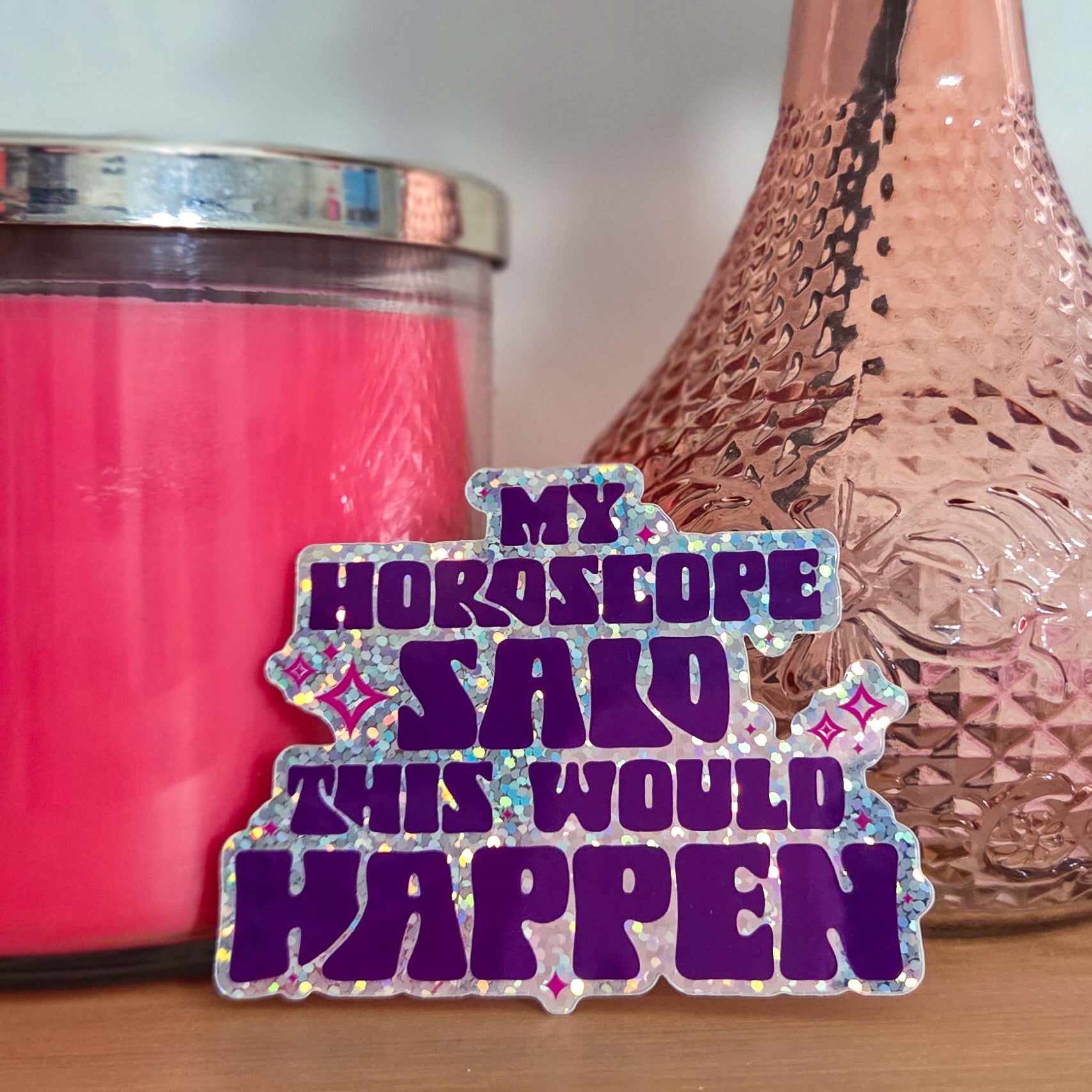 Pink candle, textured vase with a rose, and a humorous horoscope sticker on a wooden surface.