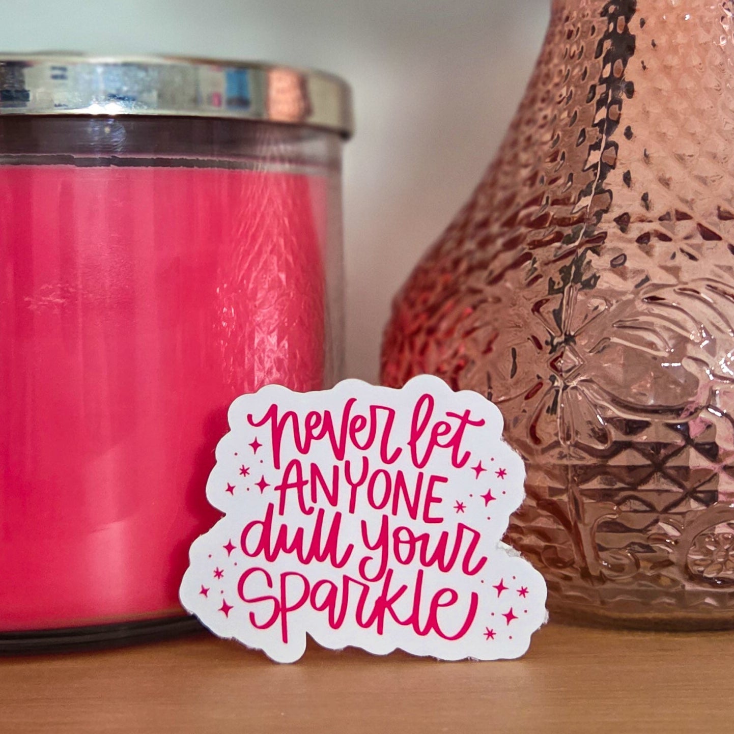 Pink candle, textured vase with a rose, and motivational sticker on a wooden surface.