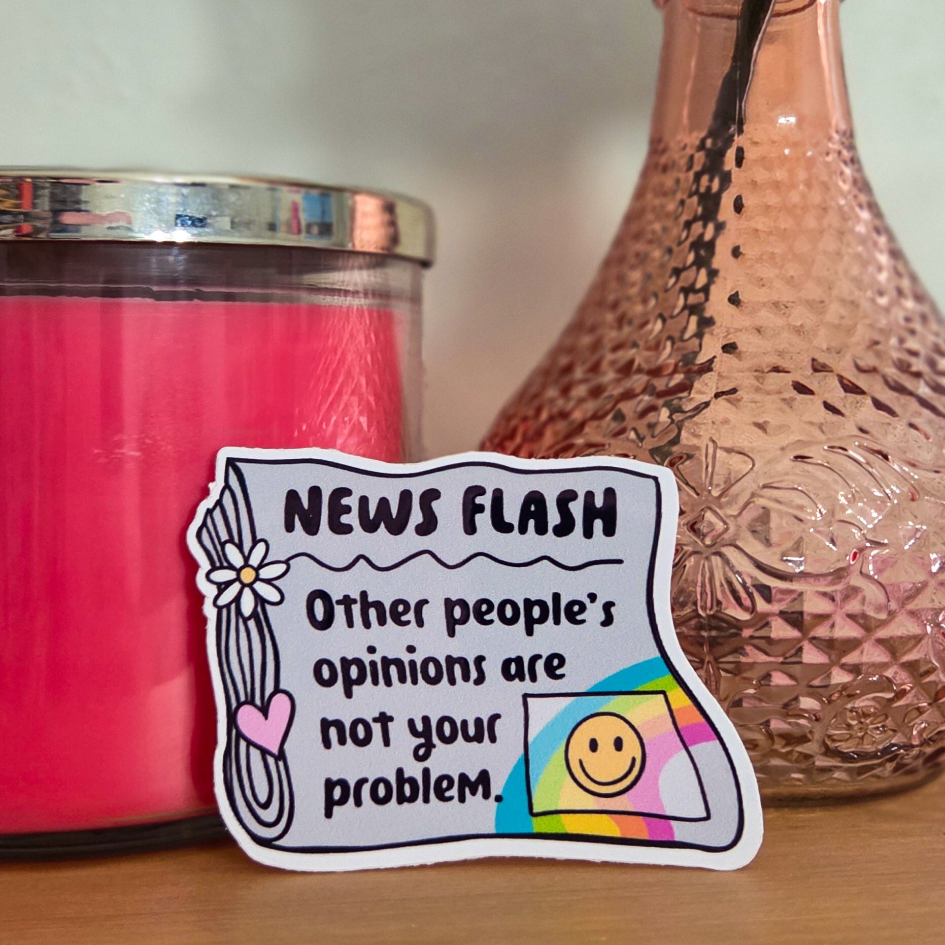 Pink candle, vase with a rose, and a colorful sticker on a wooden surface.