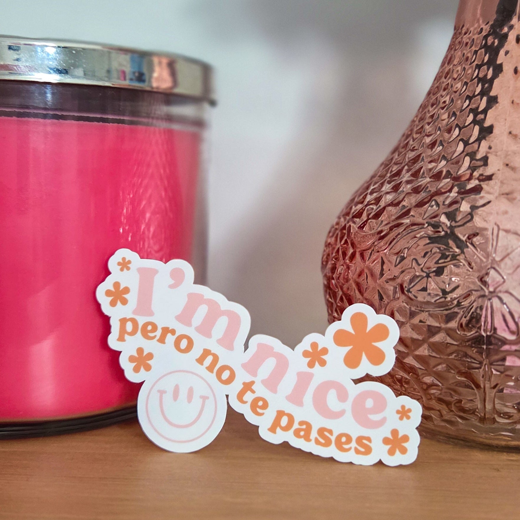 Pink candle, decorative vase and 'I'm nice pero no te pases' sticker on a wooden surface.