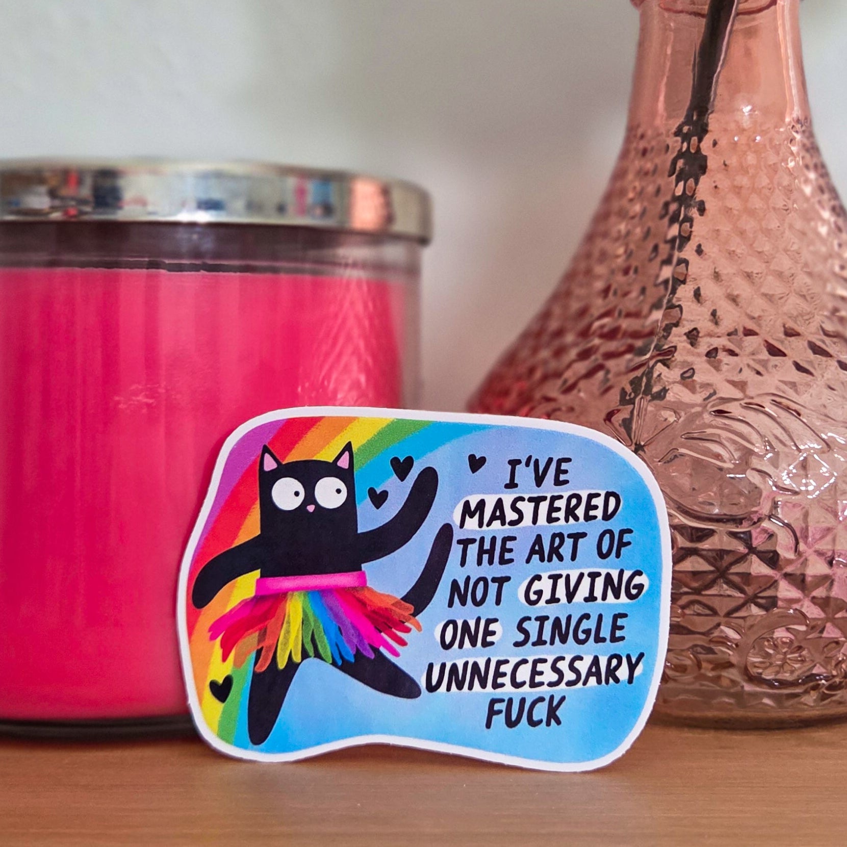 Pink candle, decorative vase with a flower, and colorful sticker on a wooden surface.