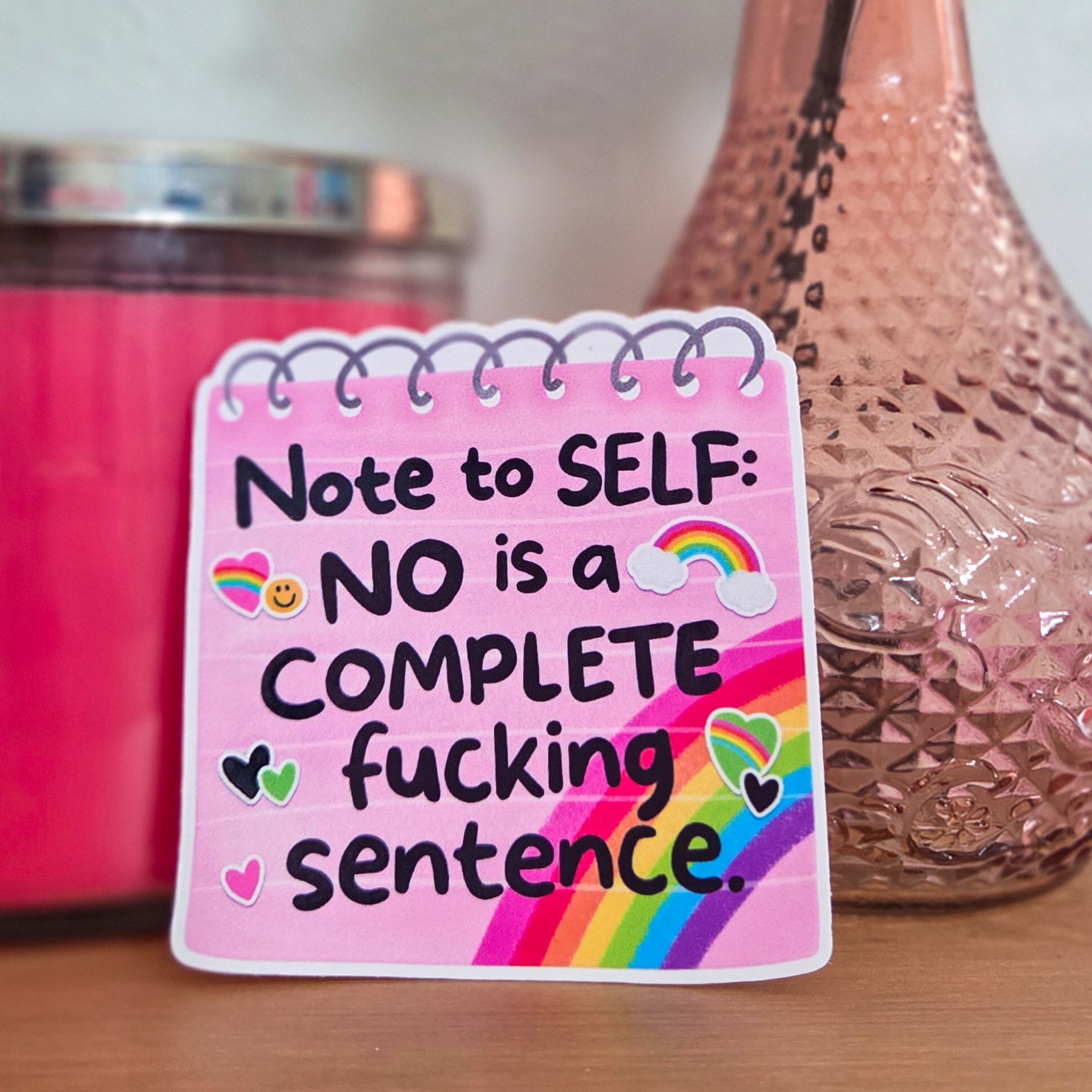 Pink notepad with motivational quote next to a vase with flowers and a candle.