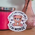 Sticker with bear graphic and text 'I'm not like other girls, I'm worse' on a wooden surface with a pink candle and vase in the background.