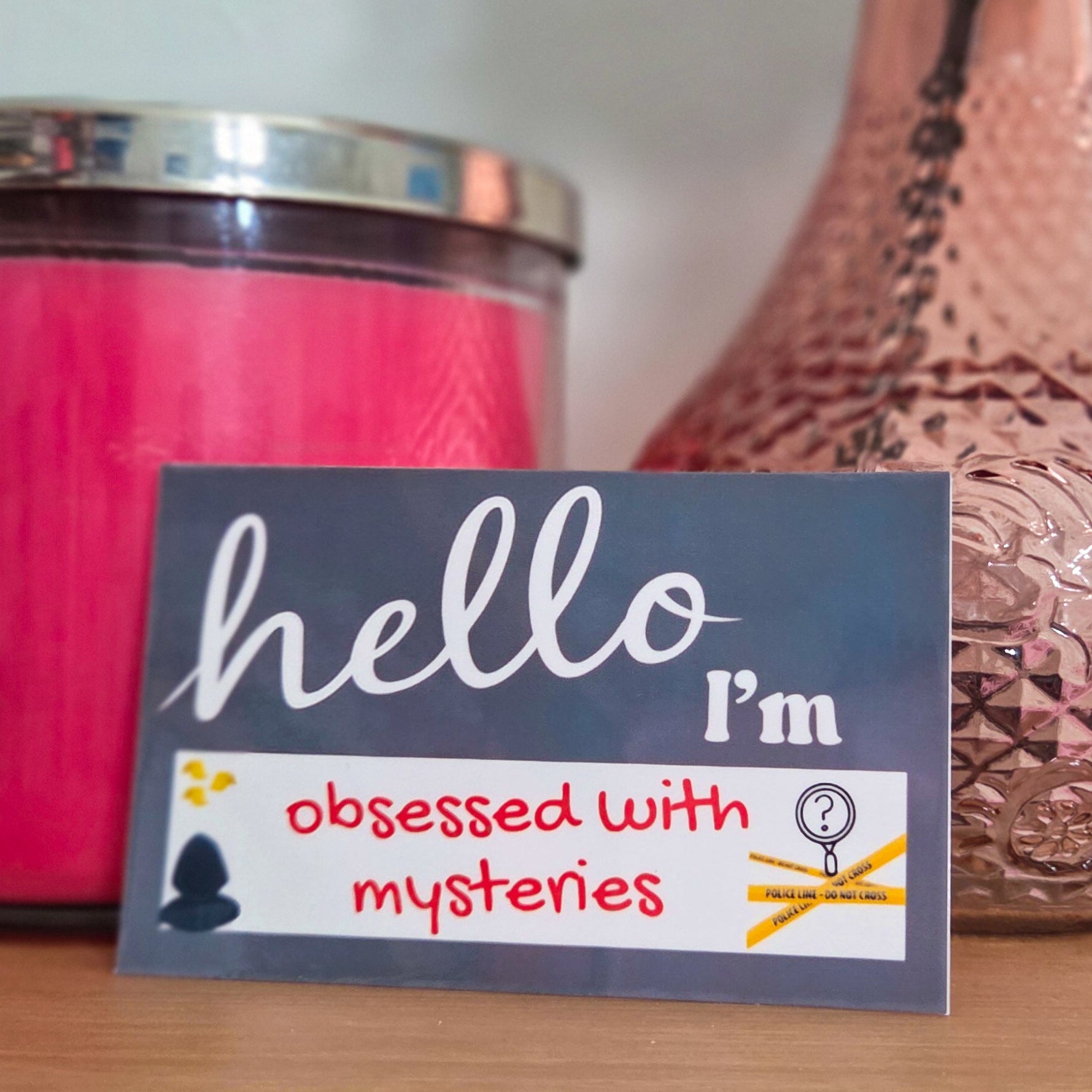 Sticker with 'hello, I'm obsessed with mysteries' text on a wooden surface with a pink candle and vase in the background.