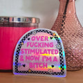 Holographic sticker with text on a wooden surface with a pink candle and vase in the background.