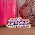 A die-cut sticker with a colorful astrological design that reads 'I'M A PISCES OF COURSE I’M TOO SOFT FOR THIS WORLD’ resting on a wooden surface.