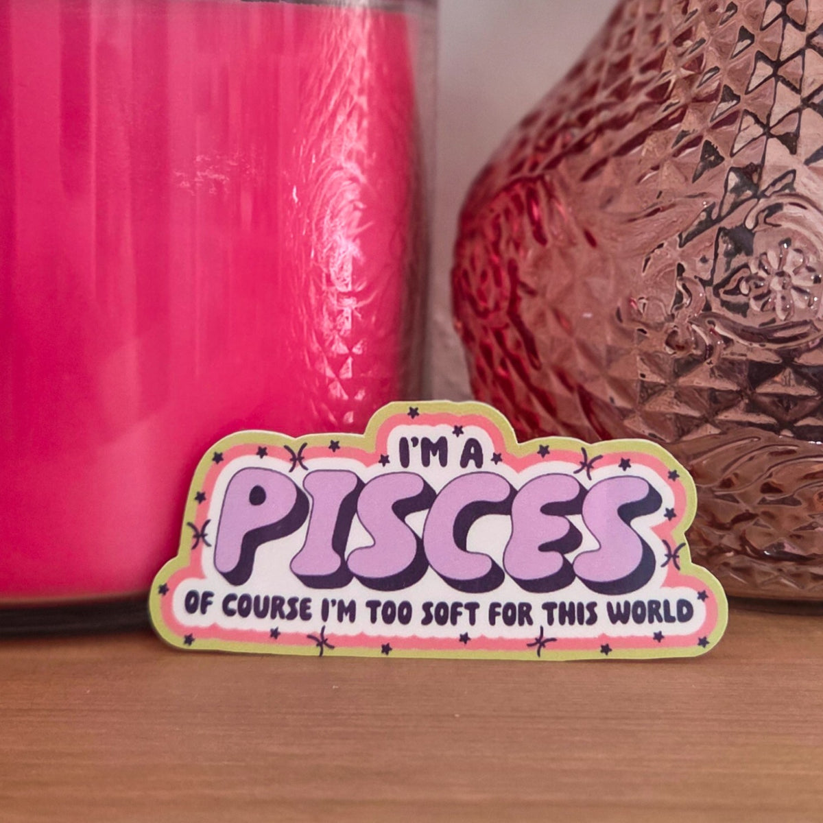 A die-cut sticker with a colorful astrological design that reads 'I'M A PISCES OF COURSE I’M TOO SOFT FOR THIS WORLD’ resting on a wooden surface.