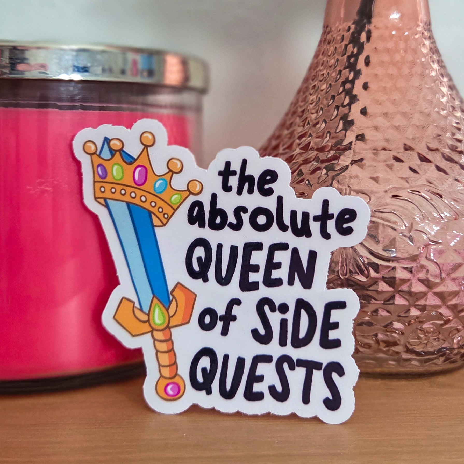 Sticker with text 'the absolute QUEEN of SIDE QUESTS' on a surface with a pink candle and vase in the background.