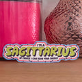 A die-cut sticker with a colorful astrological design that reads 'I'M A SAGITTARIUS OF COURSE I CAN READ YOUR ENERGY’  resting on a wooden surface.