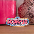 A die-cut sticker with a colorful astrological design that reads 'I'M A SCORPIO OF COURSE I’M INTENSE AF’ resting on a wooden surface.