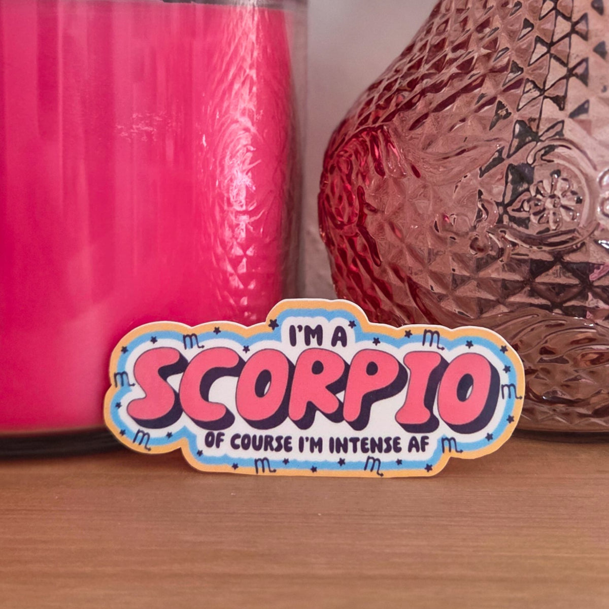 A die-cut sticker with a colorful astrological design that reads 'I'M A SCORPIO OF COURSE I’M INTENSE AF’ resting on a wooden surface.
