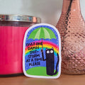 Sticker with a colorful design featuring a cat and rainbow, placed on a wooden surface next to a pink candle and vase with flowers.