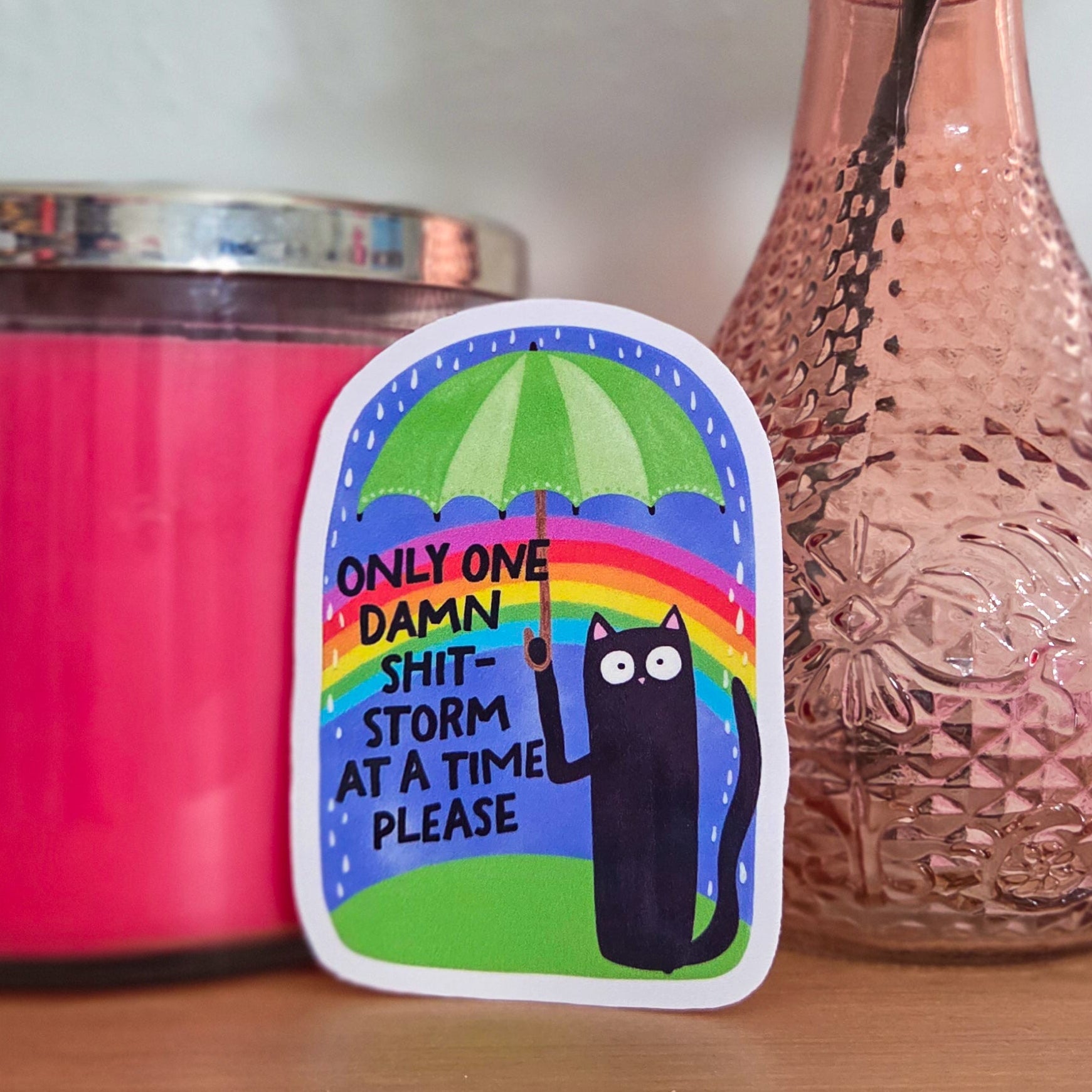 Sticker with a colorful design featuring a cat and rainbow, placed on a wooden surface next to a pink candle and vase with flowers.