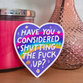 Heart-shaped sign with offensive text next to a pink candle and vase with flowers on a wooden surface.