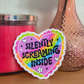 Heart-shaped sticker with 'Silently Screaming Inside' text on a wooden surface with a pink candle and vase in the background.