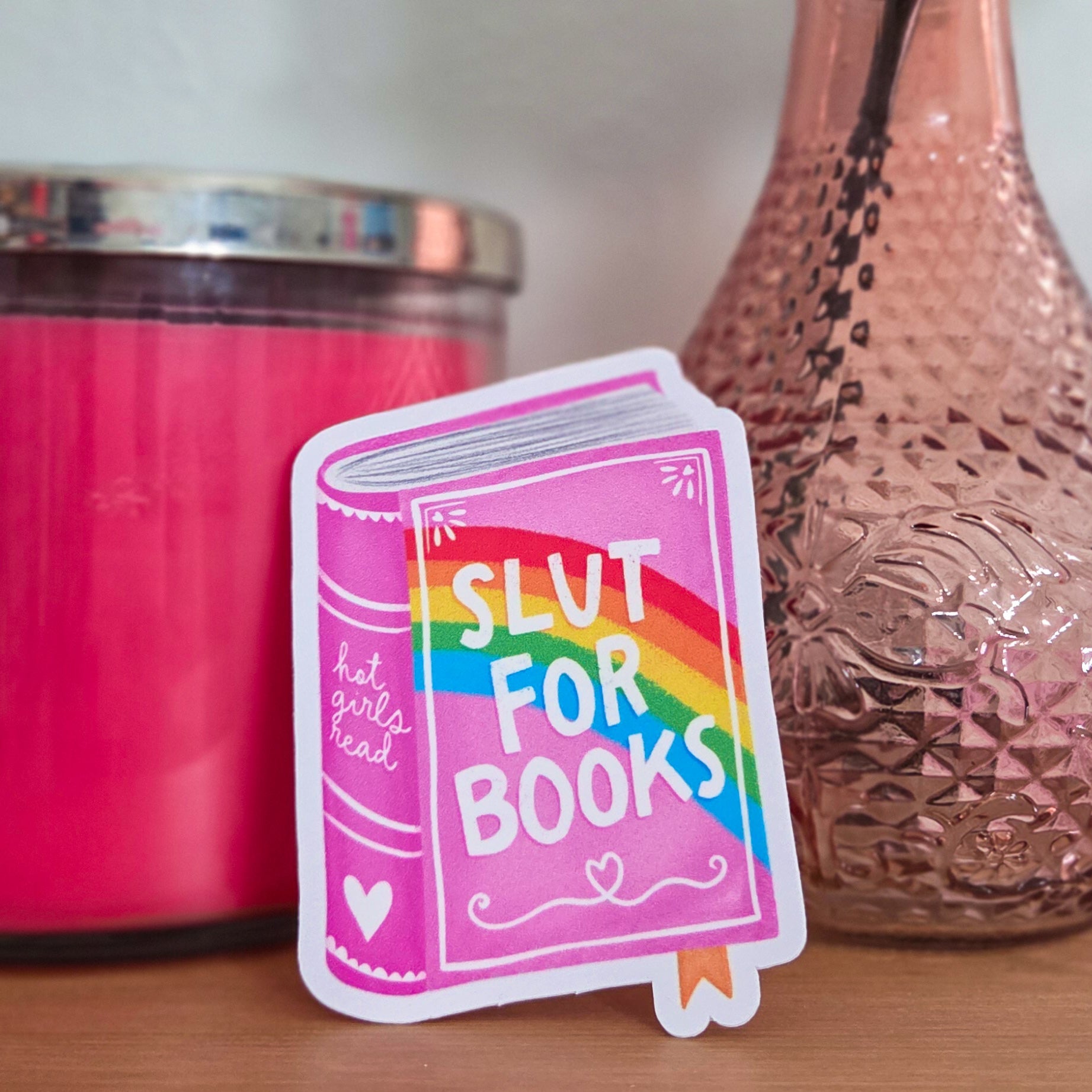 Sticker with rainbow design and text next to a pink candle and vase on a wooden surface.