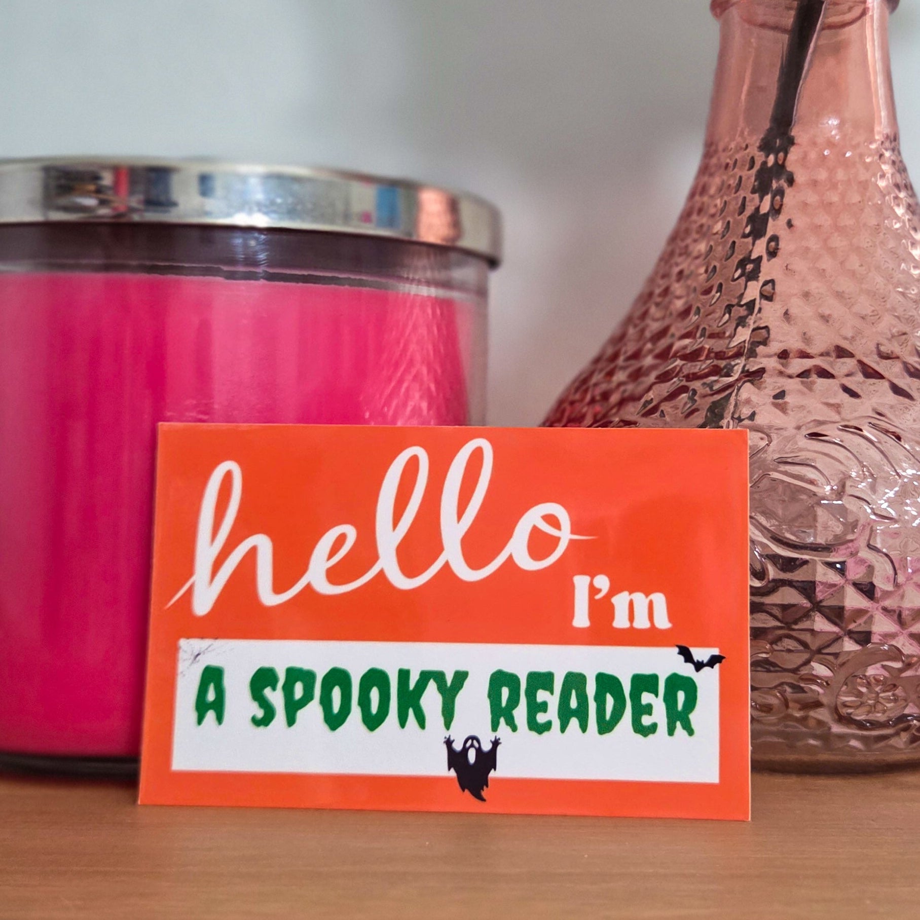 Sticker with 'hello I'm a spooky reader' text on a table with a pink candle and vase.