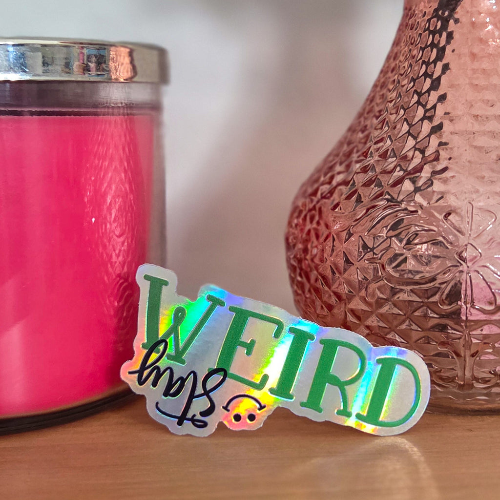 Sticker with 'Stay WEIRD' text on a wooden surface next to a pink candle and vase.