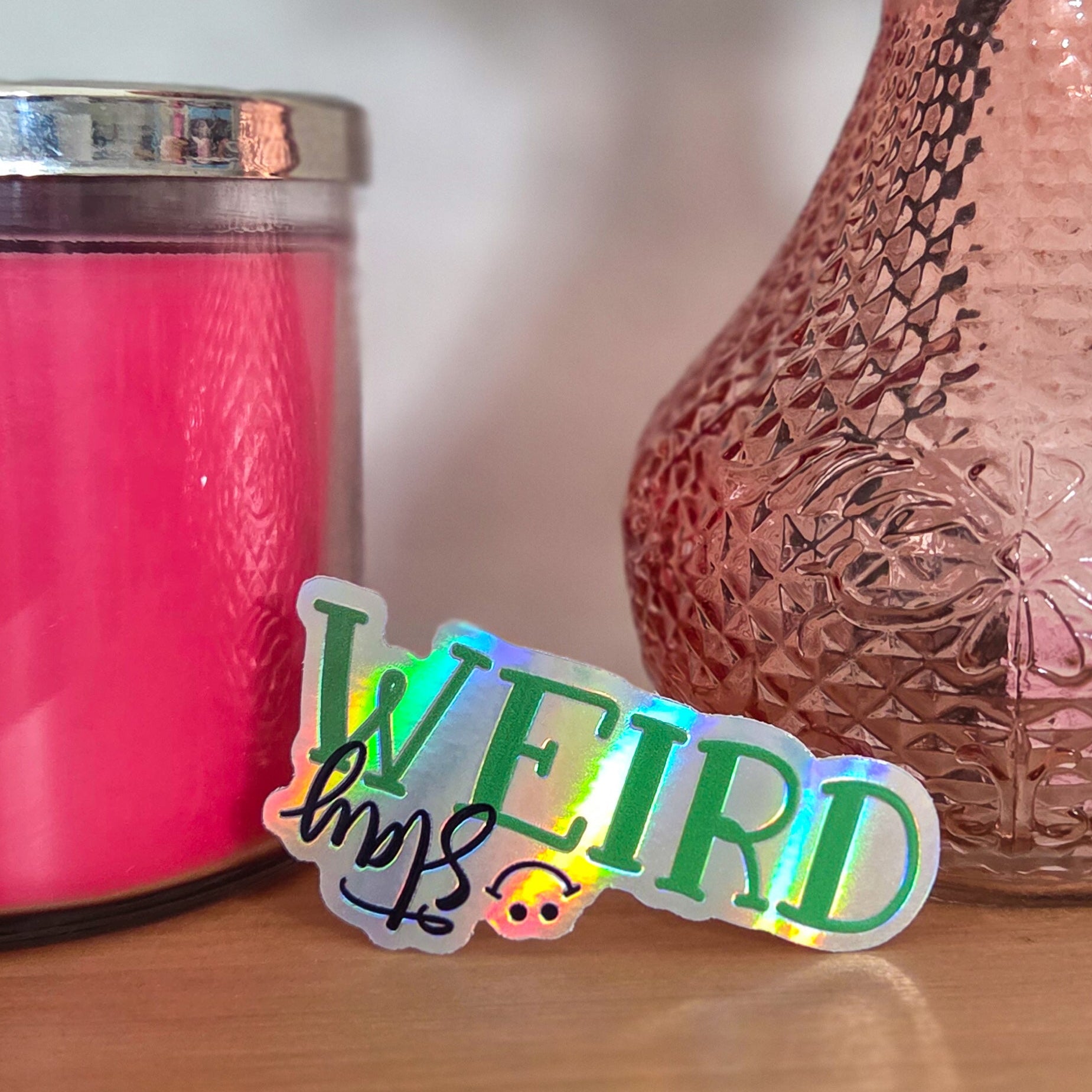 Sticker with 'Stay WEIRD' text on a wooden surface next to a pink candle and vase.