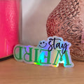 Holographic 'Stay Weird' sticker on a wooden surface with a pink vase and candle in the background.