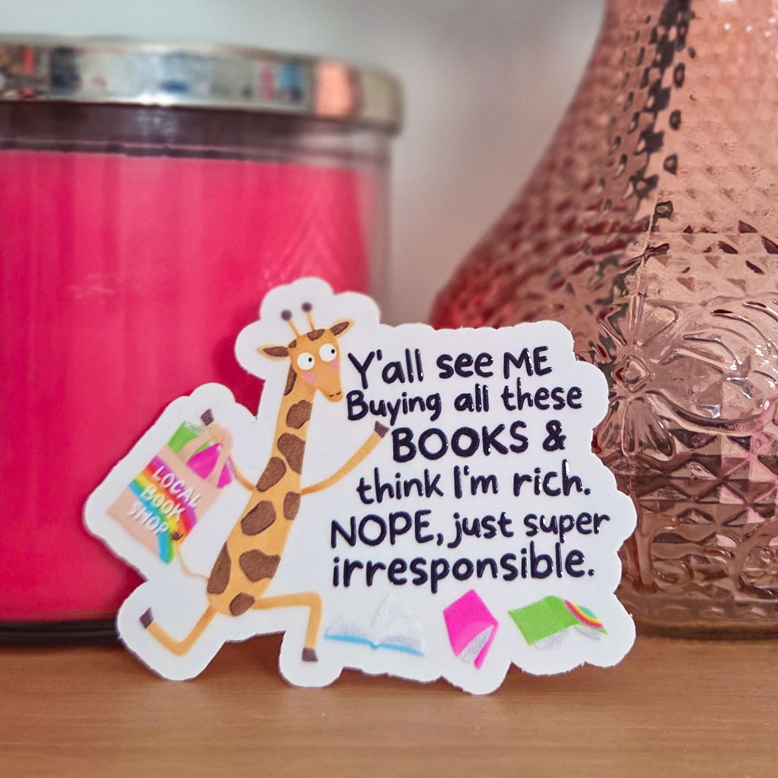 Sticker with text and giraffe graphic on a wooden surface with a pink candle and vase in the background.
