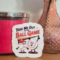 Sticker with cartoon characters and text 'Take Me Out to the Ball Game' on a wooden surface with a pink candle and vase in the background.