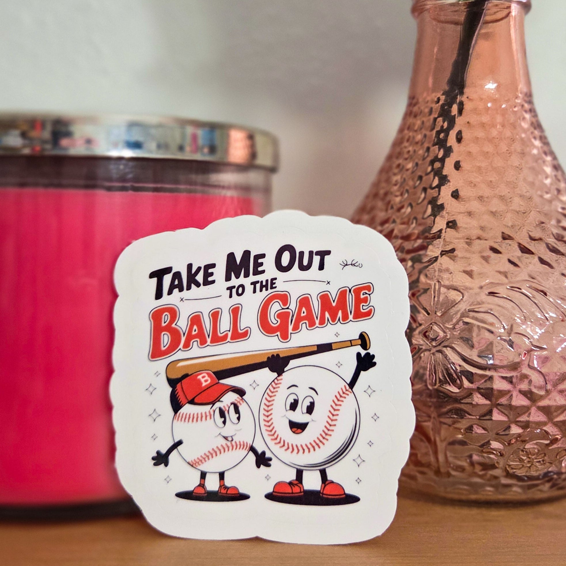 Sticker with cartoon characters and text 'Take Me Out to the Ball Game' on a wooden surface with a pink candle and vase in the background.