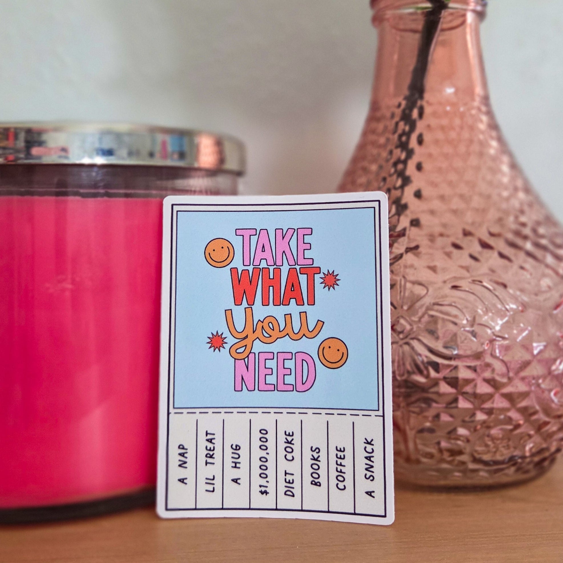 Card with 'Take What You Need' text next to a pink candle and vase with flowers on a wooden surface.