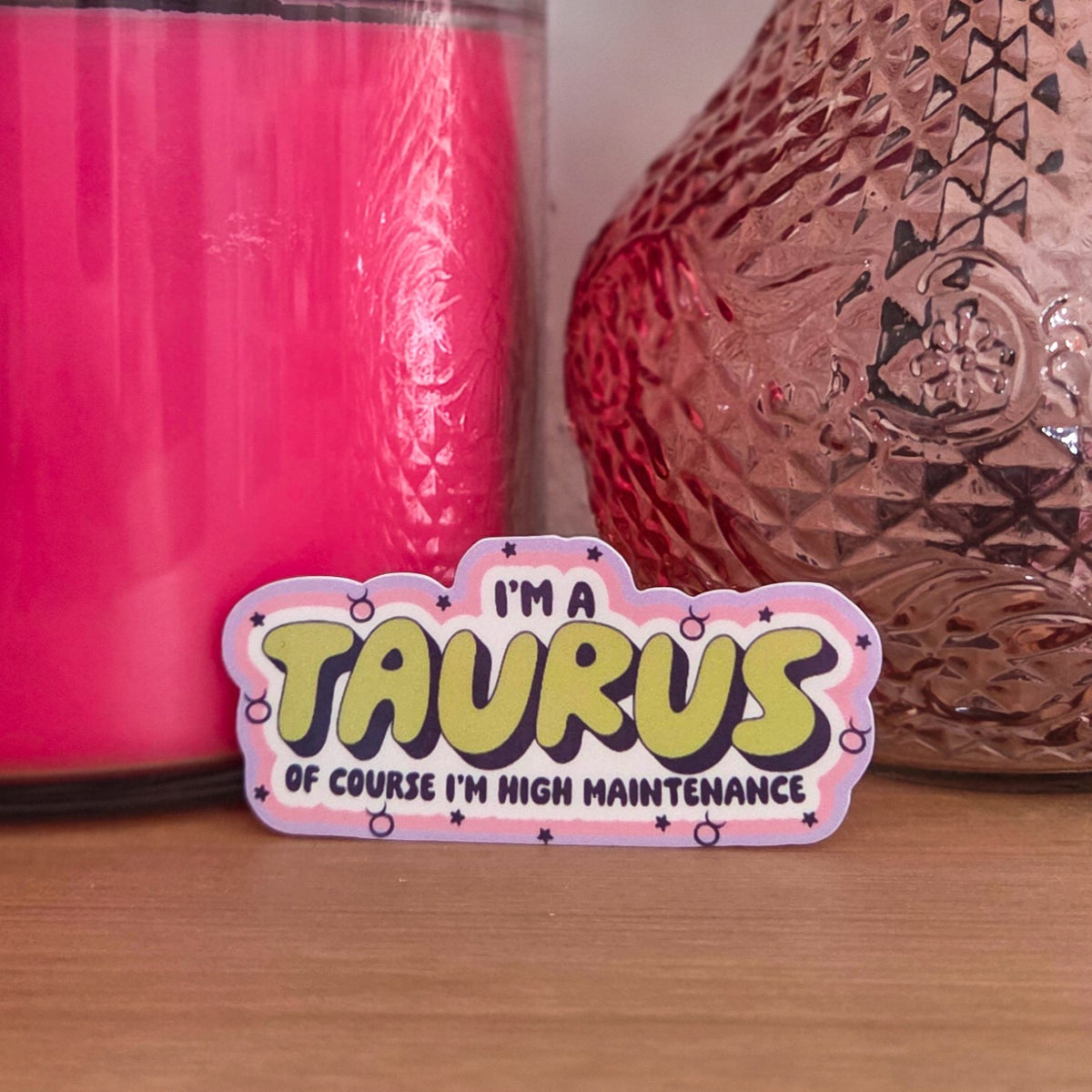 A die-cut sticker with a colorful astrological design that reads 'I'M A TAURUS OF COURSE I’M HIGH MAINTENANCE’ resting on a wooden surface.