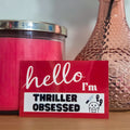 Pink candle, textured vase with a flower, and 'hello I'm thriller obsessed' sticker on a wooden surface.