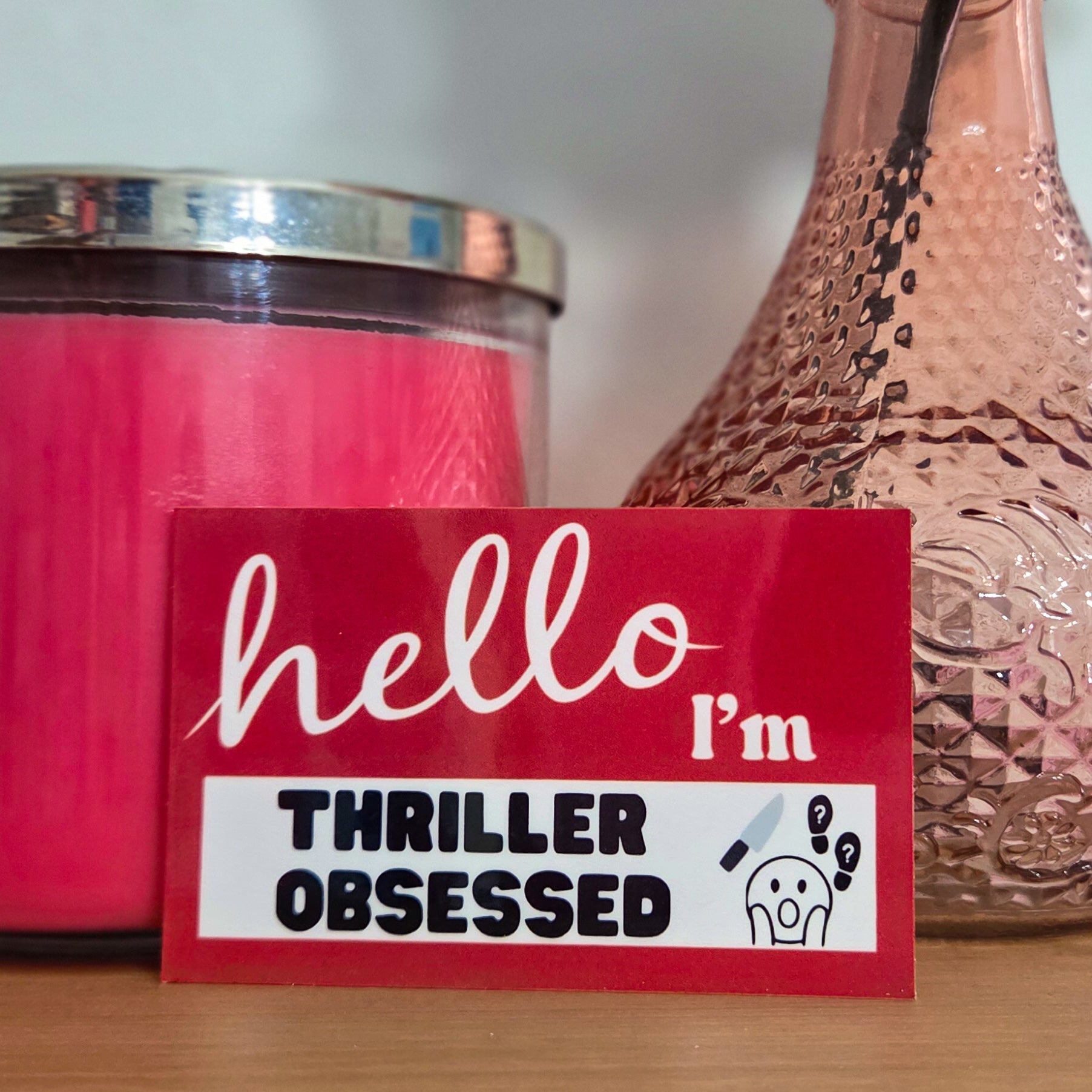 Pink candle, textured vase with a flower, and 'hello I'm thriller obsessed' sticker on a wooden surface.