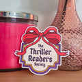 Pink candle and textured vase with a rose on a wooden surface, featuring 'The Thriller Readers Society' sticker.