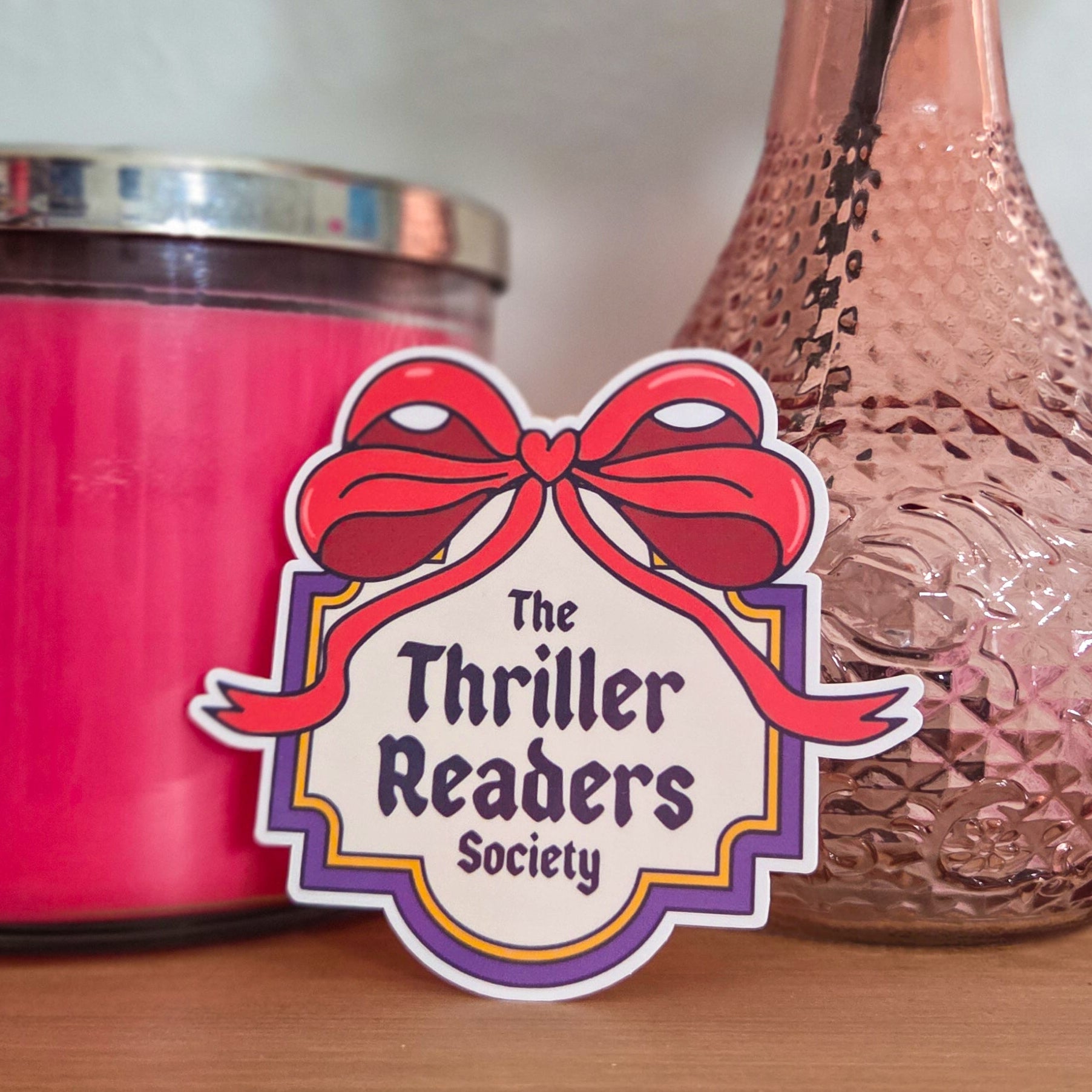 Pink candle and textured vase with a rose on a wooden surface, featuring 'The Thriller Readers Society' sticker.